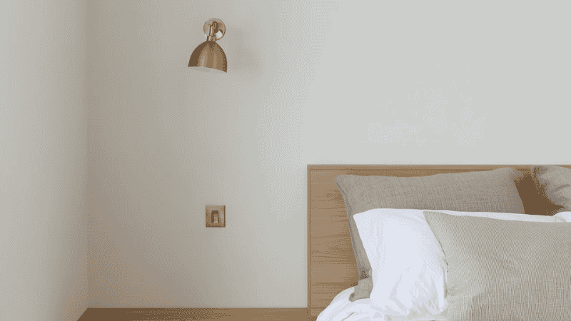 A minimalist small bedroom featuring a low-profile light oak platform bed, neutral linens, and a wall-mounted brass sconce to maximize floor space and visual flow.