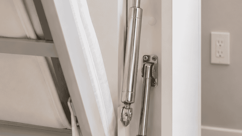 Close-up of a dual gas-piston lift mechanism on the side of a murphy bed frame, showing the chrome pistons that assist in lowering and raising the bed with minimal effort