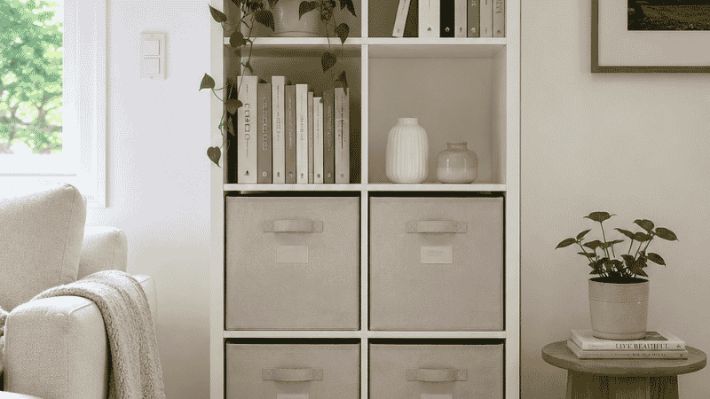 A freestanding 2x4 white cube shelving unit against a living room wall requiring no wall anchoring, lower cubes filled with opaque fabric storage bins and upper cubes displaying books and plants
