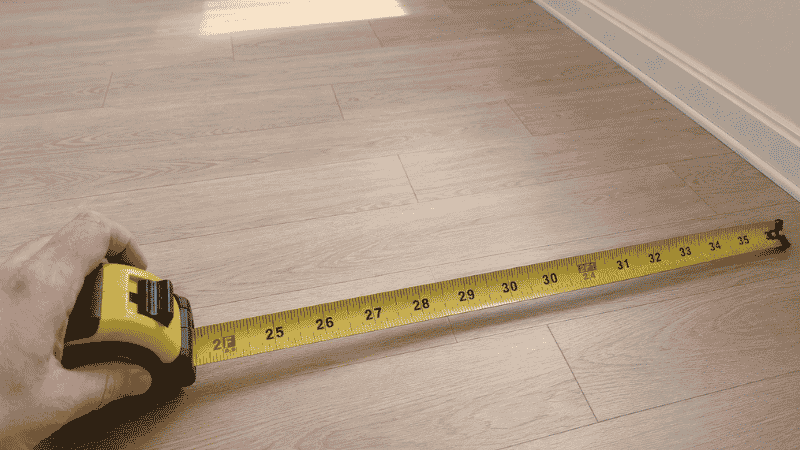 Hand holding a yellow tape measure stretched across an empty small bedroom floor before furniture placement