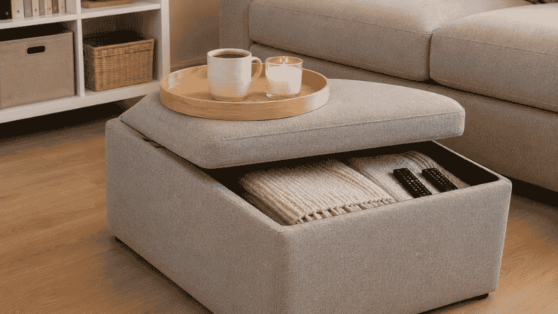 A 32-inch square light gray storage ottoman with wooden tray on top holding a coffee cup and candle, lid partially lifted on one side showing folded throws and remotes inside, functioning as coffee table in a small living room

