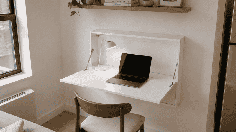 A wall-mounted fold-down desk 20 inches deep open in a studio apartment corner creating a compact work zone with a task lamp and laptop, zero floor footprint when closed
