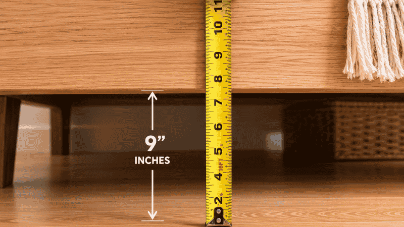 A tape measure held against the side of a bed frame measuring the clearance from the floor to the bottom rail, showing the 7-inch minimum needed for under-bed rolling storage bins
