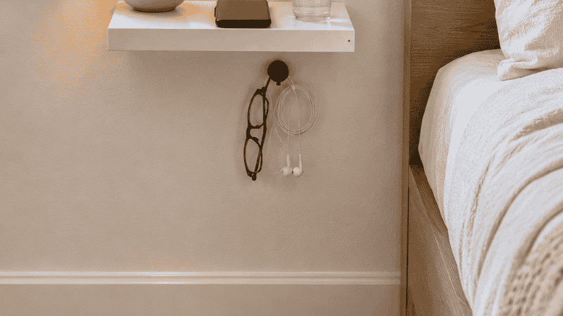 A wall-mounted floating shelf at nightstand height 22 inches from the floor beside a bed, holding a small lamp phone and glass of water with clear floor space beneath where a traditional nightstand would have stood

