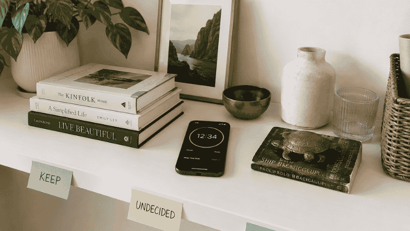 A phone timer set to 15 minutes placed beside a single shelf being decluttered, a small donate pile and keep pile visible on either side, showing the focused single-zone timed declutter method
