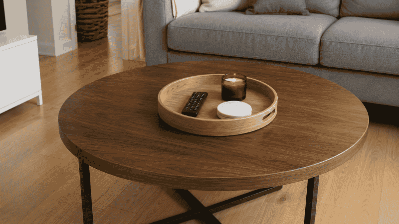 A small living room coffee table with one round wooden tray holding only a remote control, a coaster, and a small candle — demonstrating the single-tray surface rule with clear space around the tray
