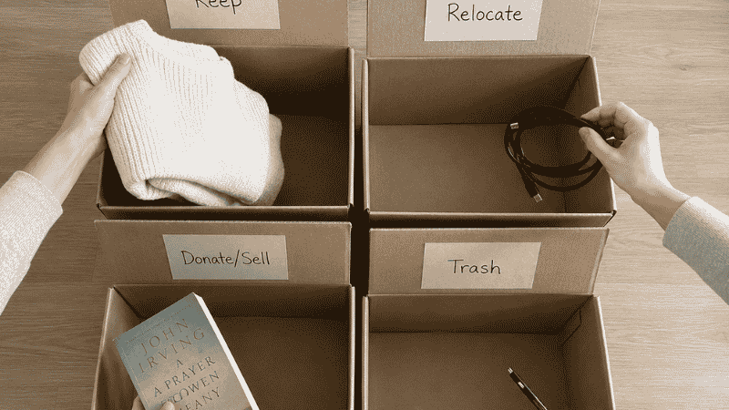 Four labeled cardboard boxes on a light floor — Keep, Relocate, Donate, Trash — with items being sorted into them during a small apartment declutter session, one shelf being emptied in the background
