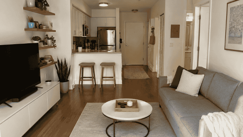 A small apartment living area shown organized and clutter-free — clear floor space, one tray on the coffee table, closed media console, and floating shelves at 70% capacity showing a functional room-by-room organization system