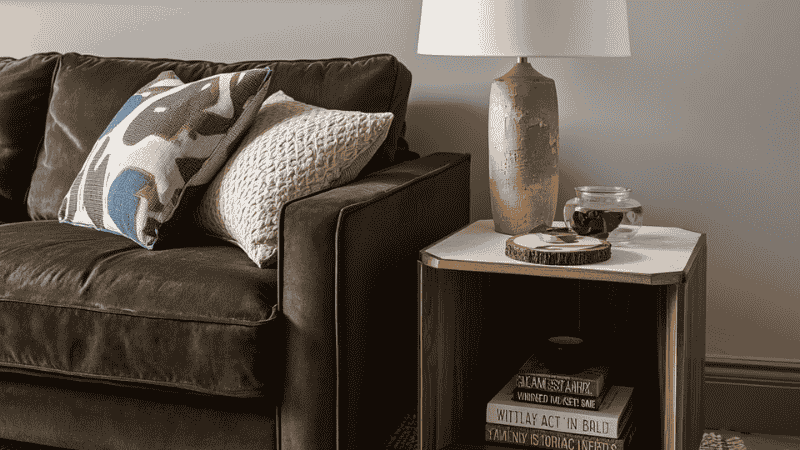 A walnut modern side table with tapered legs styled next to a low-profile grey velvet sofa in a bright, minimalist living room.