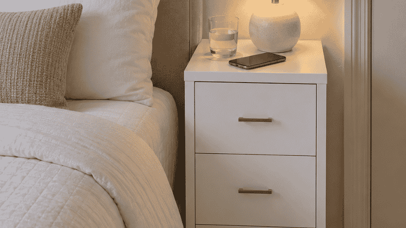 A narrow 20-inch dresser 30 inches tall positioned beside a platform bed at matching height, functioning as a nightstand with a lamp and phone on its surface and clothing stored in three drawers below