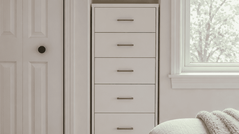 A 28-inch wide narrow dresser with 5 drawers fitting perfectly in the gap between a closet door frame and a window in a small bedroom, showing a slim white chest filling a constrained wall space