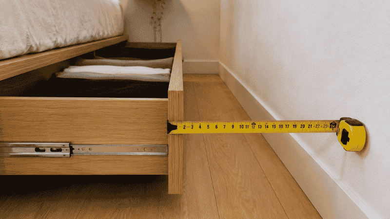 A tape measure showing 24 inches of clear floor space between an open platform bed side drawer and the bedroom wall, demonstrating the minimum clearance needed for comfortable drawer access
