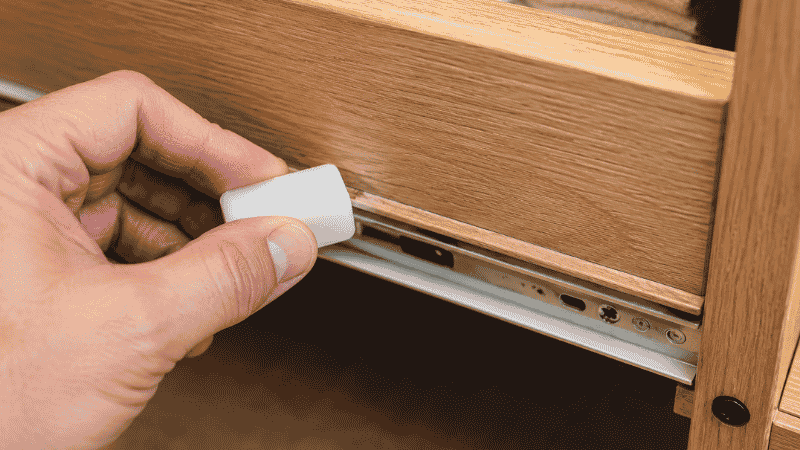 A hand rubbing a white candle wax block along the wooden runner track of a platform bed drawer, showing the DIY maintenance fix for stiff drawers that improves glide without replacement
