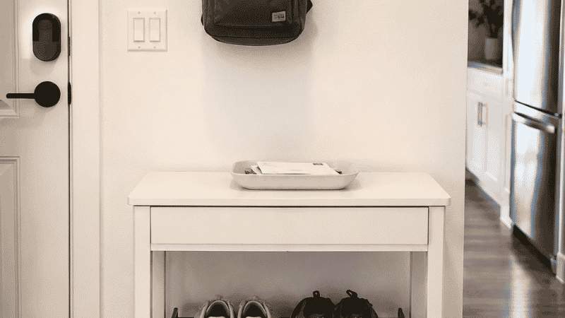 A small apartment entryway with a wall-mounted hook panel at 62 inches height holding keys on one hook and a bag on another, a small tray below for mail, and a two-tier shoe rack on the floor — showing a complete entry interception system
