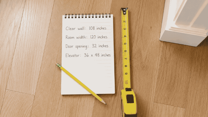 A notepad showing four written measurements — clear wall length, room width, doorway width, and elevator dimensions — beside a yellow tape measure on a light hardwood apartment floor, the essential pre-shopping checklist
