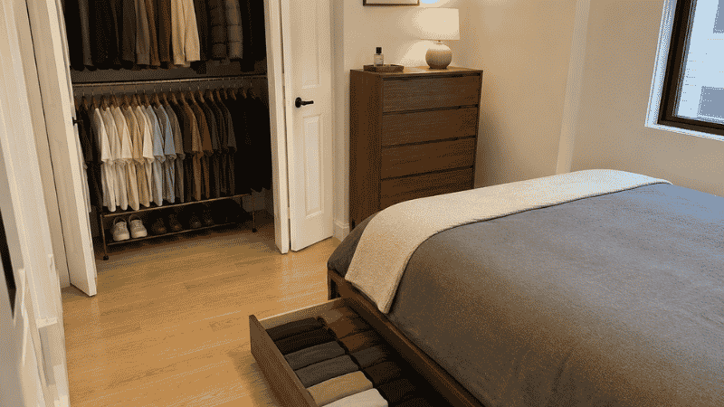 A small bedroom showing three clothing storage layers: platform bed with drawer open showing folded clothes, a double hanging rod in the open closet with shirts on the lower rod, and a slim 36-inch dresser against the wall
