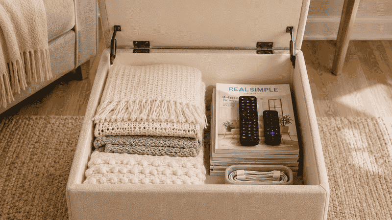 A square storage ottoman with lid fully open showing the interior organized with three folded throw blankets, a stack of magazines, two remote controls, and a small cable organizer — demonstrating real storage capacity