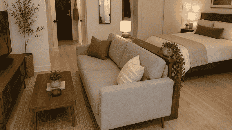 A studio apartment with a 76-inch compact sofa positioned with its back facing the sleeping zone 5 inches off the wall, a slim console table on the back side serving as a room divider, clearly separating the living and sleeping zones