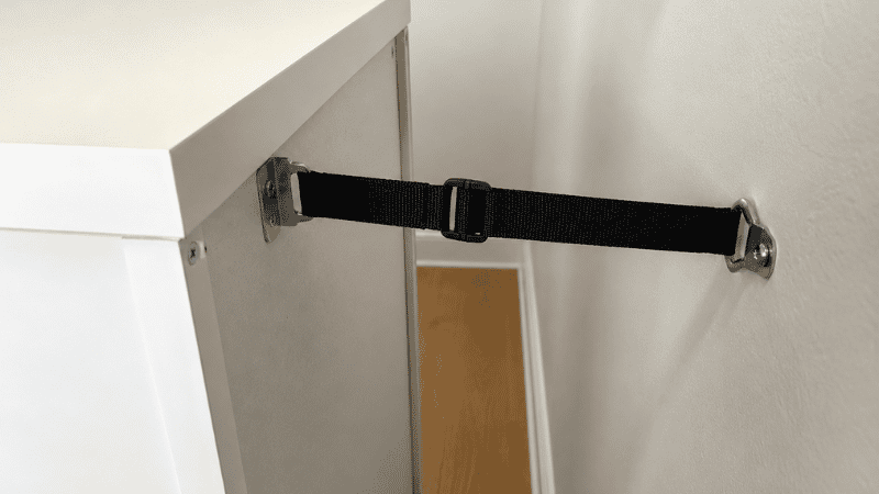 Close-up of a wall anchor strap attached from the top back of a tall narrow dresser to a wall stud, showing the anti-tip safety hardware that prevents furniture tip-over accidents