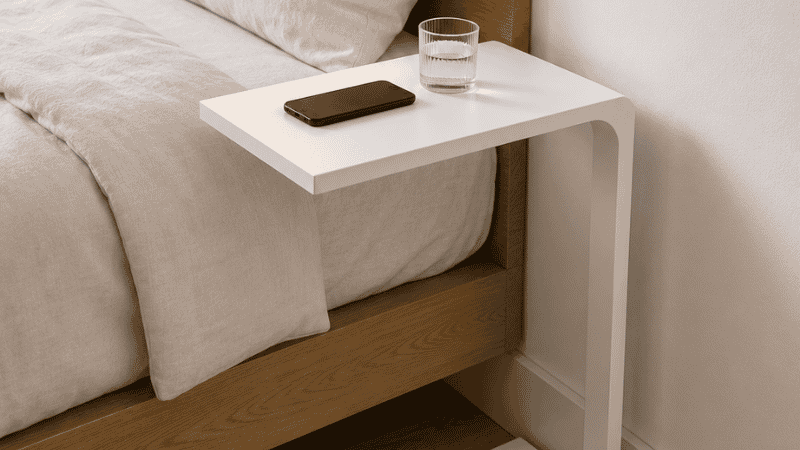 A C-shaped side table with its tabletop sliding over the mattress edge at nightstand height, the vertical support standing on the floor outside the bed frame, solving the bedside surface problem when the bed is against the wall with no gap
