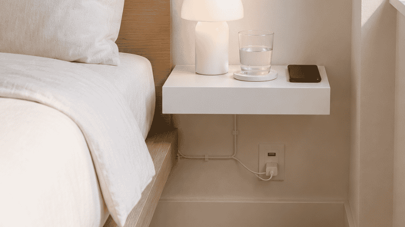 A small bedroom with a wall-mounted floating shelf at 14 inches height serving as a nightstand, with a rechargeable lamp, glass, and phone on it, the floor gap beside the bed completely clear showing the zero-footprint advantage

