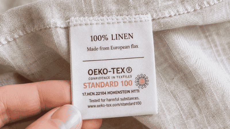 A close-up of a fabric care label on a sheet set showing an OEKO-TEX Standard 100 certification mark, indicating the fabric has been tested for harmful substances — shown as a key quality indicator when shopping for bamboo or cooling sheets
